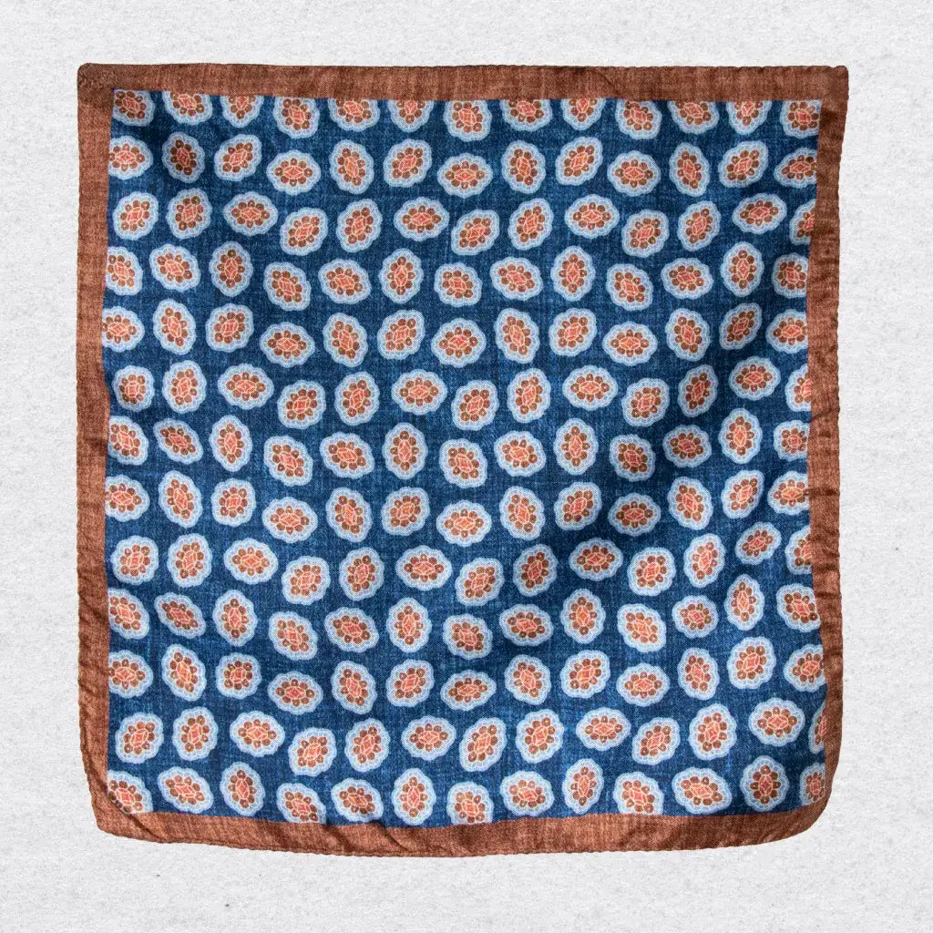 Silk pocket square with cinnamon coloured medallions and a bold border hanging against a light grey background