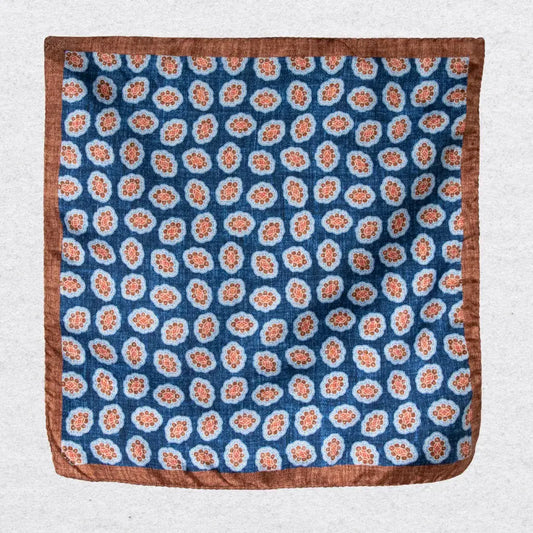 Silk pocket square with cinnamon coloured medallions and a bold border hanging against a light grey background