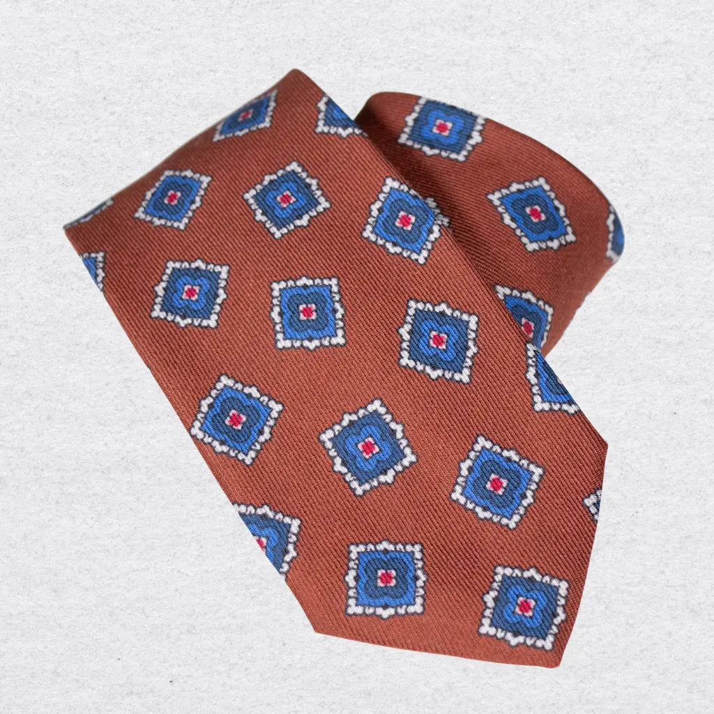 Terracotta coloured tie with blue geometric shapes, rolled on a white background