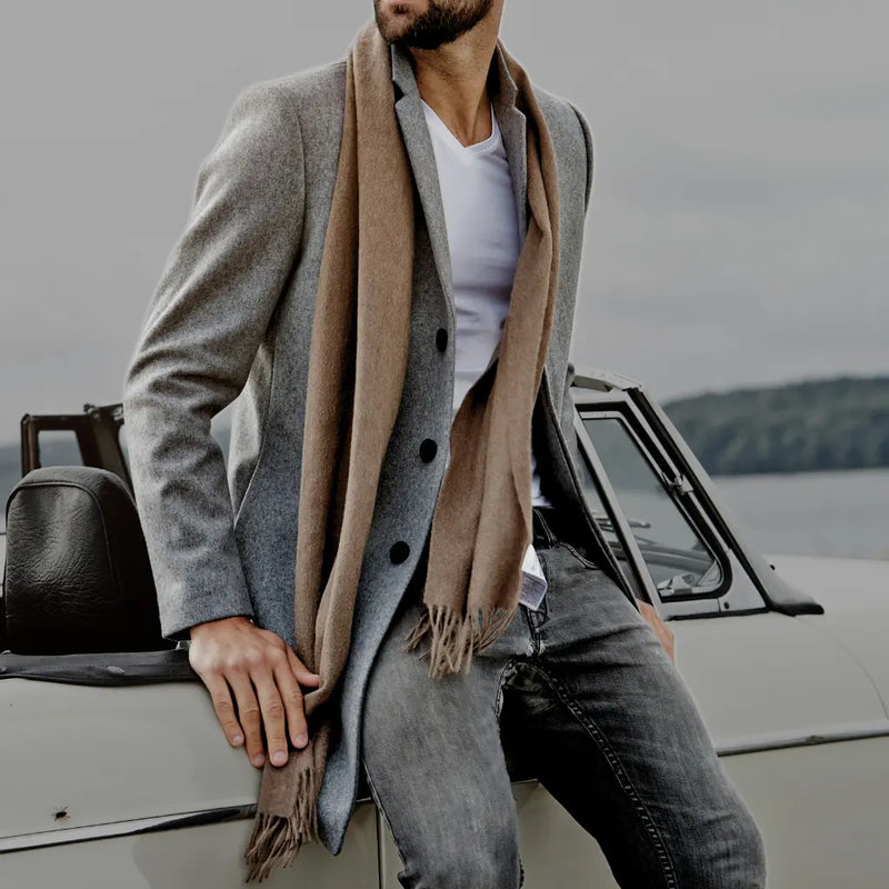 Man in a grey coat and taupe cashmere scarf standing next to a car by a lake.