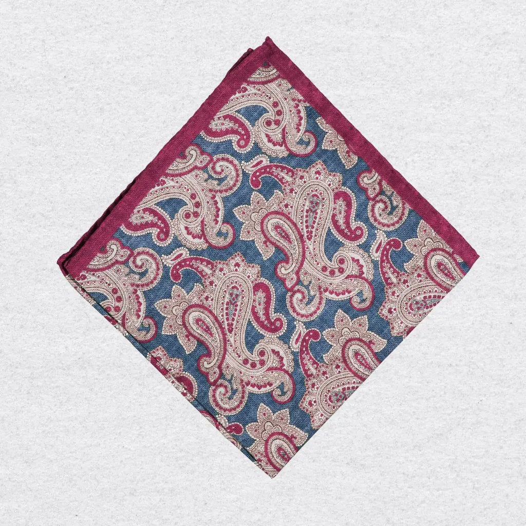Red, blue, and beige paisley patterned silk pocket square, folded on a grey background