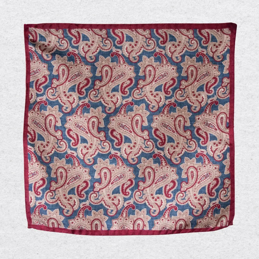 Red, blue, and beige paisley patterned silk pocket square, hanging against a light grey background